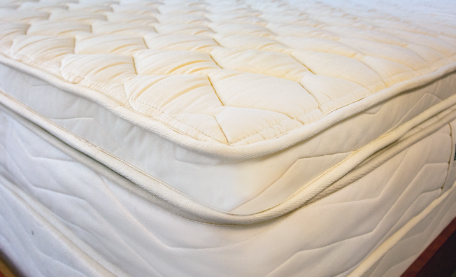 Close-up of a quilted mattress topper or mattress corner in a warm off-white/cream color, showing the stitched diamond pattern and fabric texture.