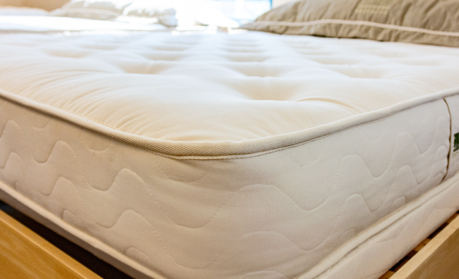 A close-up of a white tufted mattress on a wooden bed frame, with a neutral pillow visible in the softly lit background.