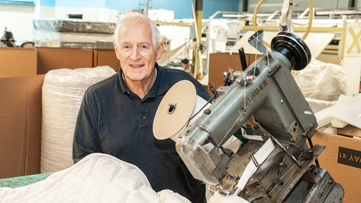 Mattress maker standing next to a sewing machine in factory.